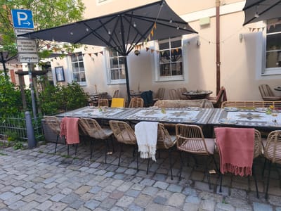 The image depicts an outdoor seating area in Erlangen, Germany, likely a restaurant or cafe, set up for dining. The foreground shows a cobblestone street, and a long table is set with plates, cutlery, and glassware. Several wicker chairs are arranged around the table, some draped with textiles. A large grey umbrella provides shade over part of the seating area, and decorative flags are strung across it. To the left, a pole with several traffic signs is visible, including a blue "P" sign indicating parking and signs with German text. The background features the facade of a building with windows. The overall scene is a neatly arranged outdoor dining space.