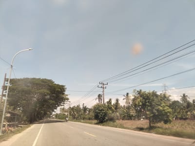 The image depicts a rural road scene in Matangkumbang, Indonesia. The road is asphalt with a yellow center line. A lone motorcycle is traveling in the distance on the left side of the road. Power lines run overhead. On either side of the road are trees, palm trees, vegetation, and occasional buildings, suggesting a tropical or semi-tropical environment. A street light stands on the left side of the road. The sky is clear and light blue.