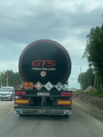 The image shows the rear view of a black tanker truck with a large black cylindrical tank. The truck is driving on a paved road that appears to be under construction or repair, with piles of dirt and gravel on the right side of the road. The truck has a license plate that reads "AT 1127 28 RUS". There are several hazard signs and identification numbers displayed on the back of the truck, including a circular sign with the number 70 inside and other diamond-shaped signs. The truck has "GTS" and "ГЛОБАЛ ТРАК СЕЙЛС" written on the tank in red lettering. There is a silver car visible in the left lane of the road. The sky is overcast. The location is specified as Radiotsentr, Russia.