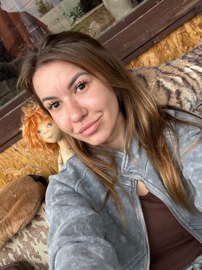A young adult female is depicted in a close-up selfie, positioned diagonally across the frame. She has long, light brown hair with lighter streaks, brown eyes, and is looking directly at the camera with a slight smile and partially opened lips. Her eyebrows are dark and well-defined, and she has prominent eyelashes. She is wearing a grey, acid-wash zippered hoodie, which is unzipped to reveal a dark brown tank top underneath. To her left, the head and upper body of a cloth doll with curly orange hair, blue eyes, and stitched facial features are visible. To the lower left, a portion of a brown, shaggy, possibly plush object is present. Behind her, a textured blanket or cushion with a striped pattern in shades of brown and purple is visible. The background consists of an oriented strand board (OSB) wall. A dark wooden beam runs horizontally behind her head, above which a window reflects an outdoor scene with foliage and what appears to be a building with a red roof. The location is Bol'shaya Mos', Russia.
