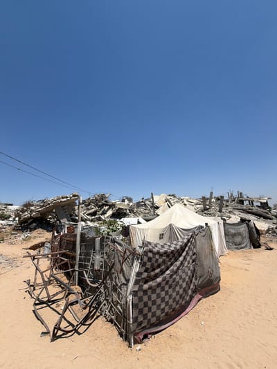 This image depicts the aftermath of destruction in Jabalia, Palestinian Territories. In the foreground, several makeshift tents are erected. One tent is partially covered by a checkered blanket, while another is draped with a patterned textile. Adjacent to the tents, twisted metal frames lie on the sandy ground. In the background, a vast expanse of rubble and debris from destroyed buildings dominates the landscape under a clear blue sky. The scene suggests a devastated urban environment with temporary shelter structures amidst the ruins.
