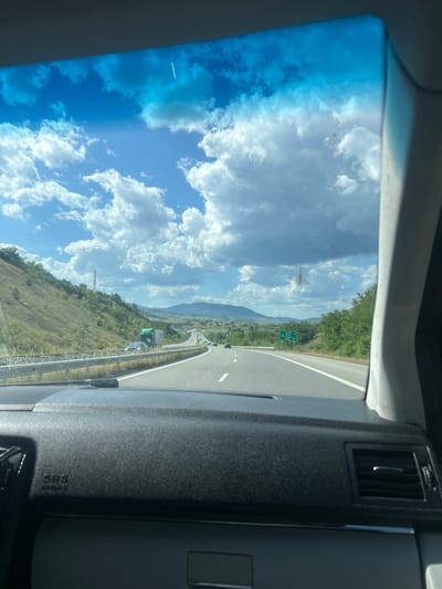 The image, taken from inside a vehicle, shows a highway landscape with a blue sky dotted with white clouds visible through the windshield. The highway has multiple lanes, and other vehicles are visible in the distance. The surrounding area includes mountains and green vegetation, suggesting a rural or mountainous setting. A cell tower is visible. A highway sign is seen in the middle ground. The car's dashboard and air vent are visible in the foreground. The location is likely near Barakovo, Bulgaria based on the presence of mountains and highways in the vicinity.