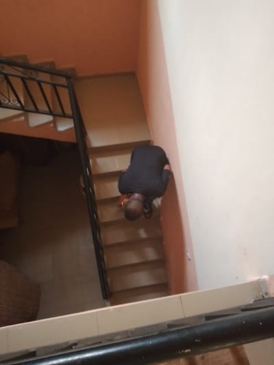 The image displays an indoor stairwell within a building in Ufuma, Nigeria, captured from an elevated perspective looking downwards. An adult male, with closely cropped dark hair, is centrally positioned on the fifth visible step from the bottom, facing the peach-colored wall. He wears a dark, long-sleeved shirt and dark trousers, adopting a crouched or kneeling posture. His hands are located near his feet, adjacent to a dark brown object consistent with footwear near his right foot. The staircase consists of light brown steps. A black metal balustrade featuring ornate scrollwork runs along the left side of the ascent. The wall to the immediate right of the staircase is off-white, while the walls framing the staircase, including the one the individual faces, are painted in a peach hue. A woven, light brown cylindrical object is partially visible on a darker floor surface in the lower-left quadrant.