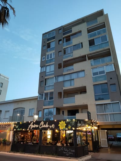 The image shows a multi-story building in Canet-en-Roussillon, France, with a restaurant or bar area at its base. The building has a modern design with balconies and many windows. The restaurant below is decorated with string lights and tropical foliage. Inside, several people are seated at tables, and some appear to be interacting with staff. The overall scene suggests a lively evening atmosphere. The street in front of the restaurant shows a few vehicles parked, and the sky is a dusky blue, indicating it is either dusk or early evening. There is a palm tree on the left side of the image.