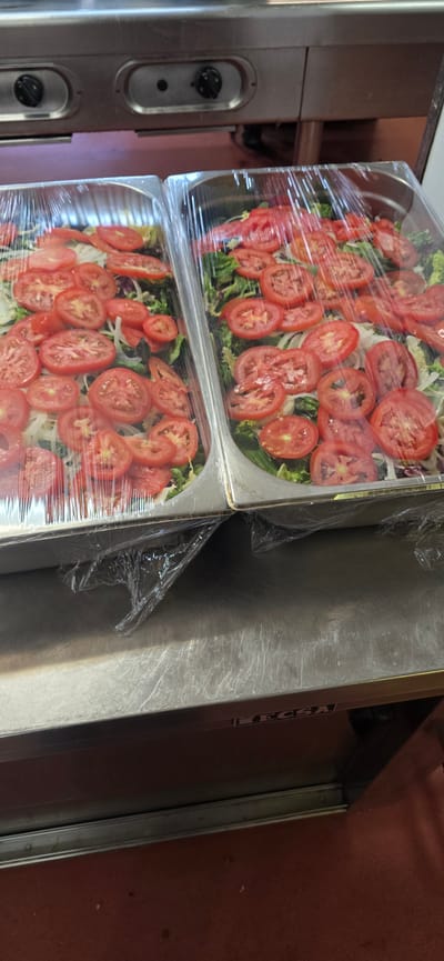 The image shows two stainless steel food pans filled with salad, covered in plastic wrap. The salads consist of lettuce, sliced onions, and numerous slices of fresh tomatoes. The food pans are stacked on top of each other and appear to be in a commercial kitchen or food service setting. To the left of the pans is a stainless steel appliance with two black knobs, likely part of kitchen equipment. To the right of the pans is a brushed stainless steel surface. The background is a reddish-brown wall. The lighting suggests it is daytime. Visible text on the side of the upper pan reads "enesa". There are no people visible in the image.