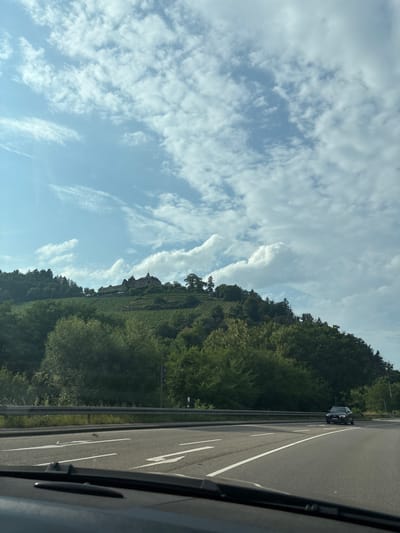 The image shows a view from inside a vehicle driving on a highway. In the distance, a hillside is covered with vineyards and trees, topped by a castle or large historic building with a tiled roof and turrets. The sky is blue with scattered white clouds. The highway has white lane markings. A dark-colored SUV is visible on the highway ahead and to the right. The foreground shows the dashboard of the vehicle. The castle appears to be located in the vicinity of Gernsbach, Germany, a town known for its vineyards and historical sites.