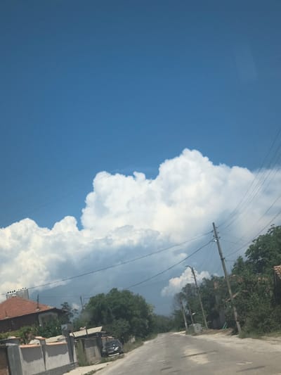 The image depicts a daytime scene in Padarsko, Bulgaria. A clear blue sky occupies a significant portion of the left frame. Dominating the right half is a large cumulus cloud formation. In the lower right corner, a section of a paved road is visible, flanked by buildings and trees lining the roadside. Utility poles with overhead lines are positioned along the road.