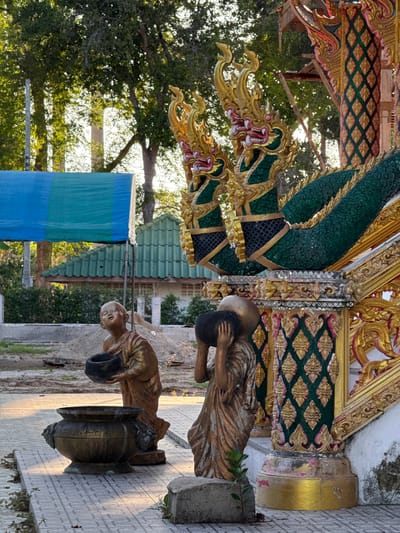 The image depicts a scene at Baan Tha Po, Thailand, featuring ornate architectural elements and statues. In the foreground, two statues of young monks are positioned on a tiled pathway. The monk on the left, facing the viewer, holds a black bowl in his hands. The monk on the right is depicted from behind, carrying a larger black object on his head, supported by his hands. A large, weathered brass pot with decorative carvings sits to the left of the statues.

Dominating the right side of the image is a grand structure adorned with vibrant, golden, dragon-like sculptures. These serpentine figures have green scales and intricate golden details on their heads and necks, with red accents around their mouths. The structure itself is elaborately decorated with gold trim and green panels featuring a diamond-shaped pattern.

Behind the statues and architectural elements, a blue and green tarp is visible, stretched between two poles. Further in the background, a green tiled roof of a building can be seen, along with lush trees that create a dappled sunlight effect. The ground appears to be a mixture of dirt and gravel in some areas. The overall impression is one of religious or cultural significance, likely within a temple or similar sacred site.
