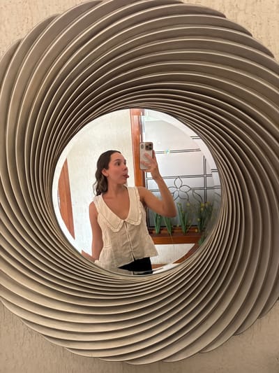 The image captures a female individual in her twenties or thirties taking a mirror selfie in an indoor setting located in Ciudad Victoria, Mexico. She is reflected in a large, circular mirror with a distinct, textured frame featuring concentric, spiraling ridges in a light, neutral tone. The woman wears a sleeveless, light-colored top with a prominent, rounded collar and a central row of fasteners, along with dark lower garments. Her right hand, adorned with a ring, holds a light-colored smartphone with multiple camera lenses and a visible magnetic attachment, positioned to capture the reflection. She is looking towards the phone/mirror with her tongue extended in a playful gesture. The environment reflected in the mirror behind her includes a textured, light-colored wall, a segment of a wooden doorframe or trim, and a window or decorative panel with a grid pattern incorporating a central floral motif. Below this, a wooden shelf holds a row of green plants interspersed with small yellow flowers, appearing to be artificial. The lighting suggests an interior space.