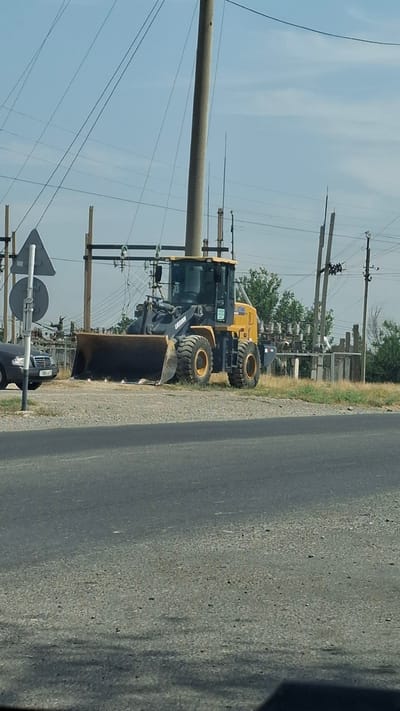 A yellow and black front-end loader is parked on a gravel surface next to a paved road. The loader has a large bucket at the front. In the background, there are utility poles with wires, electrical insulators, and a line of trees. To the left of the loader, a dark-colored car is parked. A traffic sign with a white triangle and a circular sign is visible to the left of the loader. The scene appears to be in an outdoor, possibly rural or semi-urban, area with clear skies. The location is identified as Қасымбек датқа, Kazakhstan.