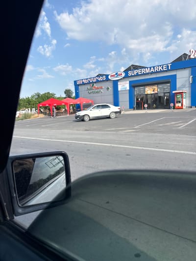 The image depicts a storefront with the sign "SUPERMARKET" in large white letters on a blue background. Below it, a logo reads "INESSA". The building has large windows displaying products. To the left of the supermarket entrance, there's a red and white sign with text in Armenian. A white car is parked in front of the building.

The scene is outdoors, with a bright blue sky and scattered white clouds, suggesting it's daytime and the weather is clear. The ground appears to be asphalt, marked with white lines, indicating a parking area or road. In the foreground, a car's side mirror is visible, reflecting trees and part of the building. There are also red tents set up near the supermarket, possibly for vendors or an outdoor event.

Visible text includes "SUPERMARKET", "INESSA", and Armenian script on a sign. A coffee machine is also visible on the building's exterior. The overall impression is of a commercial area on a sunny day.