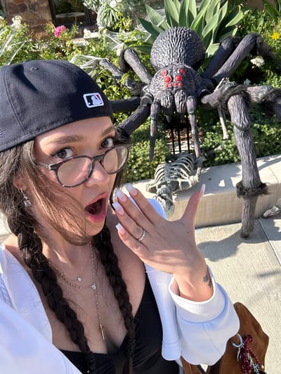 This image captures an individual posing for a selfie outdoors in Manhattan Beach, United States. The person, wearing a black baseball cap with a white MLB logo and patterned eyeglasses, has dark hair styled in two braids. Their mouth is open in a surprised or exclamatory posture. The left hand is raised, prominently displaying a ring with multiple small stones on the ring finger and long, square-tipped nails with a white-to-pink ombre manicure. Multiple gold-toned necklaces, including a delicate chain with a small heart charm and a longer rosary-style chain with a cross pendant, are visible. The individual wears a white long-sleeved top or jacket over a dark inner garment.

In the background, a large, detailed black prop spider with red eyes and a textured body stands prominently amidst green foliage. Beneath the spider, a white skeleton prop lies on the ground. Artificial white cobwebs are draped over the surrounding plants and shrubs, indicating Halloween decorations. A stone-clad building facade is partially visible in the upper left background. The scene is illuminated by bright, clear daylight, suggesting a residential front yard setting.