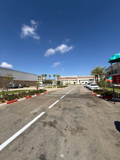 A low-angle shot looks down a two-lane paved road in an industrial or commercial district under a clear blue sky with scattered white clouds. The road is marked with solid white lines and has red and white curbs lining both sides. On the left, a long, grey, corrugated metal building stands behind a green lawn and some small trees. On the right, a series of modern buildings with light-colored facades are visible, interspersed with palm trees and green landscaping. Several cars are parked along the right side of the road, including white sedans and silver SUVs. A bright green canopy structure is visible on the far right, along with a pole with colorful elements, suggesting a playground or recreational area. The overall scene depicts a well-maintained outdoor area with a mix of industrial and commercial architecture. The location is the Reishit 10 complex in Pardes Hana-Karkur, Israel.