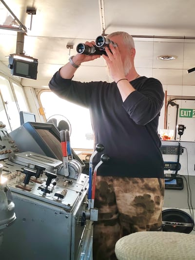 The image shows a man in a ship's cabin looking through binoculars. The man has short gray hair and a gray beard. He is wearing a black long-sleeved shirt and camouflage patterned shorts. He is standing in front of a control panel with various levers and knobs. Behind him is a white wall with an "EXIT" sign in green lettering. There is radio equipment on the wall. The scene appears to be on a ship, possibly a ferry or a commercial vessel, in a brightly lit interior. There is no specific information in the image to confirm the location as Anisimovo, Russia.
