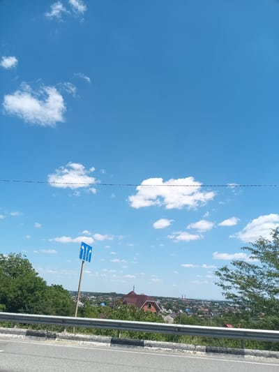 A bright, sunny day is captured in this vertical, medium-shot photograph taken from a roadside perspective, overlooking the city of Krymsk, Russia. The sky is a vibrant, clear blue, dotted with fluffy white cumulus clouds. In the mid-ground, a blue traffic sign with three white arrows indicating lanes for either going straight or turning left is prominently displayed on a pole. Behind the sign, a dense, green, treed landscape unfolds, leading to a residential area with houses, some with red roofs, nestled amongst more trees. A few industrial structures, possibly factories with tall smokestacks, are visible in the far distance, suggesting the presence of industry on the outskirts of the city. A metal guardrail runs along the bottom of the frame, separating the road from the embankment, and the asphalt road surface is visible in the foreground. The overall impression is of a typical summer day in a Russian town.