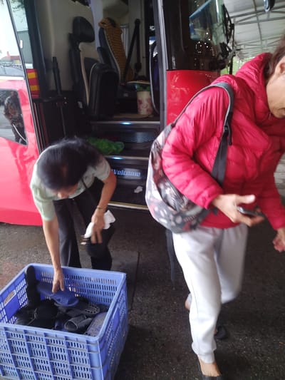 In Chu Văn An Ward, Vietnam, the image shows two people next to a red bus with its door open. One person, wearing a light green top and dark pants, is bent over a blue plastic crate filled with dark-colored sandals, holding a white cloth and a sandal. The other person, wearing a red puffer jacket and white pants, with a gray shoulder bag, is walking away from the bus, looking down and holding a cell phone. The bus has "Welcome" written on the step. The ground is dark and appears to be concrete.