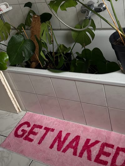 This image depicts a bathroom interior in Bad Nauheim, Germany. The primary subject is a white tiled bathtub, filled with multiple potted plants, predominantly large-leafed *Monstera* varieties, some with visible fenestrations and supported by a coir moss pole. Water droplets are present on the plants' leaves and the rim of the bathtub, suggesting recent watering. Above the bathtub, a wall-mounted chrome faucet with an attached flexible shower hose is visible against a light-colored tiled wall. The floor is also tiled with light-colored square tiles. A pink rectangular bath mat, bearing the phrase "GET NAKED" in darker pink capital letters, lies on the floor in front of the bathtub. A section of a white door frame or trim is visible along the left edge of the image, resting on the floor tiles. No individuals are present in the scene.
