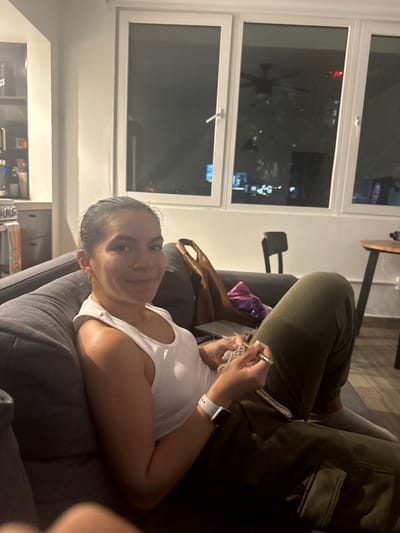 A woman in a white tank top rests on a gray sofa, holding knitting needles and yarn. Another person, dressed in olive green pants, is beside her. A large window illuminates the room, reflecting city lights. In the background, a small round table is visible. The woman faces the camera.