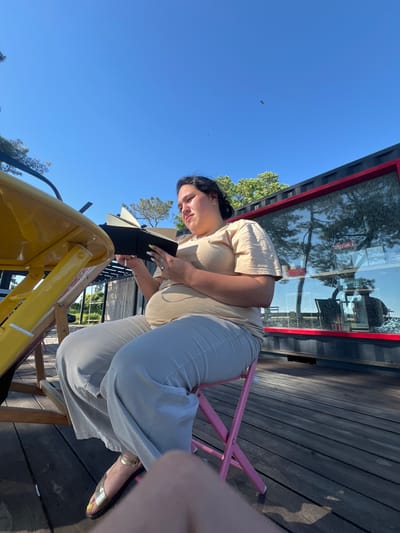 In this media file, a woman is captured from a slightly elevated perspective. She is seated on a pink chair, her body turned to her left as she looks down at a black notebook resting on a yellow easel. She holds a pen in her right hand, poised as if to write or draw, and her left hand is positioned on the notebook. She is wearing a beige, short-sleeved top and light-colored pants, with gold sandals on her feet. Her hair is dark and pulled back.

The setting appears to be outdoors, with a clear blue sky above and wooden planks forming the ground. Behind the woman, a building with a large glass window framed by a red border is visible. Reflections of trees and what appears to be a "Coca-Cola" logo can be seen in the glass. The presence of the easel and notebook suggests an activity like sketching or journaling. The lighting indicates it is daytime, likely sunny, given the clear sky and the shadows cast. The overall mood seems relaxed and focused, with the woman engrossed in her activity. The location context provided is Ochamchire, Abkhazia.