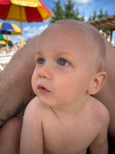 A close-up, tilted shot focuses on the face of a young child with light hair and striking blue eyes. The child is held by an adult, whose arm is visible in the foreground. The child's skin has a few droplets of water, suggesting they may have been swimming or playing in water. They have a neutral expression, with their gaze directed slightly away from the camera.

In the background, out of focus, are colorful elements that resemble parts of a playground or amusement area. There are large, umbrella-like structures with bright red, yellow, and blue sections. The sky is visible, appearing to be a clear blue with some white clouds. The overall setting seems to be outdoors, possibly at a water park or a recreational area on a sunny day.

The image is oriented such that the child's head is at the top right, and the adult's arm and the background elements extend towards the bottom left. There is no visible text in the image. The time of day appears to be daytime, based on the bright lighting and clear sky.