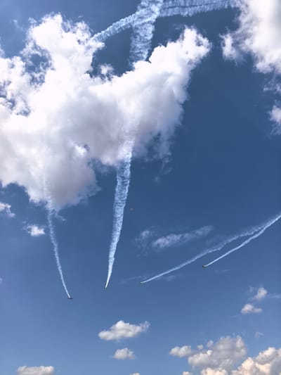 The image captures an aerial performance against a bright blue sky with scattered white clouds. Several planes are visible, leaving trails of white smoke. Some of these trails form intricate patterns, suggesting aerobatic maneuvers. One small, distant plane is colored orange and white. The overall scene is clear and detailed, suggesting it was taken on a sunny day. The specific location in Nestiukovo, Russia, is not visually discernible from the image itself.
