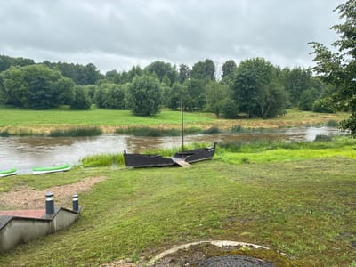 The image depicts a cloudy day scene along a riverbank in Sabile, Latvia. In the foreground, a grassy slope leads down to a river. A dark wooden boat, appearing to be a replica of a historical vessel with a mast, rests on the grass near the water's edge. Two green and white kayaks are also visible on the bank further to the left. In the lower left of the image, there is a partial view of a structure with a red tiled roof and two modern-looking lights. The river itself is brown and choppy, with tall green reeds lining its banks. The background is filled with a dense forest of various green trees. The overall atmosphere is one of a peaceful, natural landscape, possibly a park or recreational area.