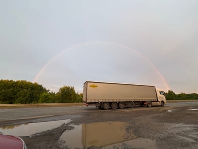 This media file depicts a roadside scene in Sovkhoznyi, Russia, under an overcast sky that shows a faint rainbow. A large, light-colored semi-truck with a tan trailer is driving on a paved road. The trailer has a yellow logo with text that is partially visible, appearing to say "ORTRAUS". The road surface is wet and shows signs of disrepair, with visible potholes filled with water and gravel. Reflecting in the puddles is the truck and the surrounding environment, including a dense line of green trees bordering the road. A metal guardrail runs along the edge of the road, separating it from the trees. The sky is a pale gray, with the subtle arc of a rainbow visible in the upper portion of the frame. The overall impression is of a cool, possibly damp, day, with the truck in motion on a journey. The time of day appears to be late afternoon or early evening, given the soft lighting and the presence of the rainbow.