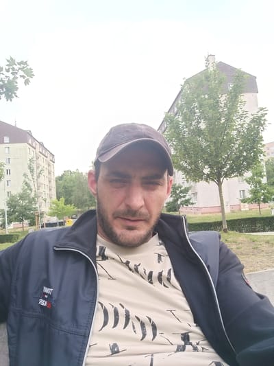 A man wearing a dark baseball cap and a dark zip-up jacket over a light-colored t-shirt with a black abstract pattern is taking a selfie outdoors. The man has a beard and is looking towards the camera. In the background, there are apartment buildings, trees, and a cloudy sky. The scene appears to be in a residential area, possibly a park or courtyard, in the city of Milovice, Czechia.