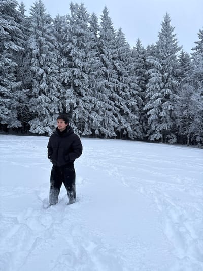A male individual stands in a snow-covered open field in Le Hohwald, France. The individual, appearing to be in his late teens or early twenties, is positioned slightly to the left of the frame's center, facing partially right with his head turned left. He is dressed in a black puffer jacket and black pants; the lower half of his pants from the knees down are visibly wet and covered in snow. His hands are placed in his jacket pockets. The ground is uniformly covered in a thick layer of white snow, showing numerous footprints and tracks in the foreground and midground, particularly to the right. Directly behind the individual and extending across the background is a dense forest of coniferous trees, each branch heavily laden with a thick coating of snow and frost. The sky above the tree line is uniformly overcast and light grey, characteristic of a winter day.