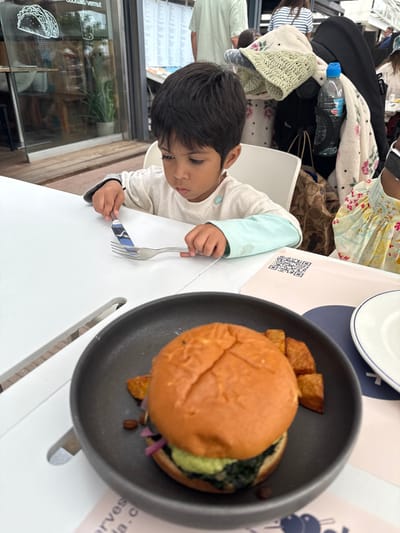 A young male child, approximately 2-4 years old, with dark hair and eyes, is seated at a white slatted table in an outdoor or semi-outdoor restaurant setting in L'estartit, Spain. He wears a light-colored long-sleeve shirt with light blue accents and holds a silver-colored fork in his left hand and a knife in his right, looking intently downward at the utensils. In the foreground, on the table to the right, a dark grey plate contains a burger with a brioche bun, green and dark green fillings, red pickled onions, and dark seeds, accompanied by several square-cut, golden-brown fried potatoes. A white plate with a blue rim is partially visible to the right of the burger plate. A light pink paper with a QR code and the partial text "harvest" and "cocktails/vermut" lies on the table. Behind the child, through a large glass partition, a sign with a taco graphic and the words "cocktails / vermut" is visible, along with reflections and indistinct background figures. A black stroller draped with a light-colored, patterned blanket and a clear plastic water bottle in a holder is positioned behind the child. Other patrons are visible in the background, including a person in a light green shirt.