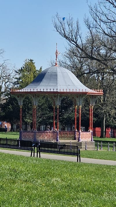The image shows a Victorian-style bandstand in a park setting. The bandstand has a domed roof, which is white with a blue accent, and ornate metalwork in shades of orange and purple decorates the structure. It is surrounded by green trees and grass. To the right, there is a black bench. The sky is clear and blue. A blue object is visible in the sky, possibly a kite or balloon. The bandstand is located in the city of Lincoln, United Kingdom.