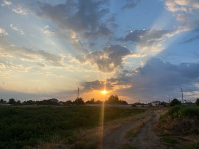 The image depicts a sunset over a rural area, likely Kurtovo Konare, Bulgaria. The sky is a mix of blue and grey clouds, with rays of sunlight piercing through, creating a dramatic effect. The sun itself is partially obscured by clouds, casting a warm golden glow on the scene. In the foreground, a dirt road winds through a field of tall dry grass and green vegetation. Scattered houses and trees are visible in the middle ground, suggesting a residential area bordering the countryside. Power lines stretch across the upper portion of the frame, indicating the presence of infrastructure. The overall atmosphere is serene and picturesque, capturing the beauty of a rural sunset.