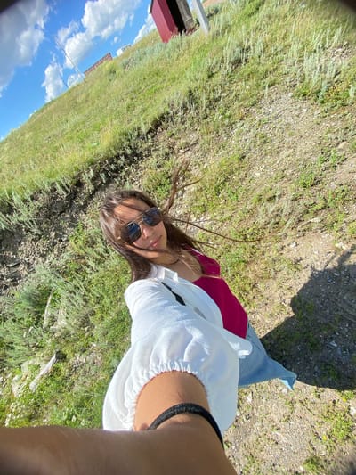 In Yuldybayevo, Russia, a young woman is captured in a selfie. She is wearing sunglasses, a white shirt, and a pink top. Her hair is dark and windswept. The background is a grassy hillside with sparse vegetation and rocky ground. A red building can be seen in the distance on the top of the hill. The sky is blue with scattered white clouds. The image is taken outdoors on a sunny day.