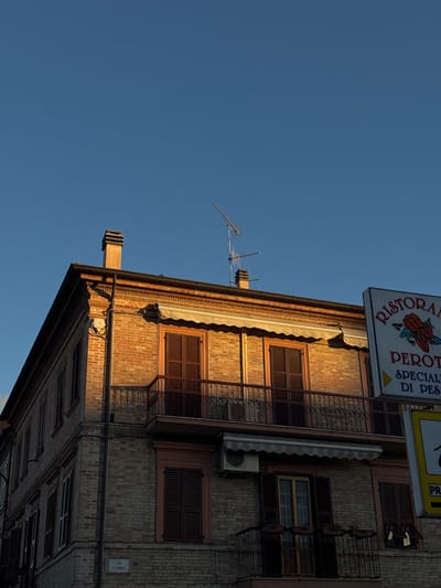 The image displays a multi-story, corner-facing brick building in Pedaso, Italy, specifically located on Via F. D'Anna, as indicated by a white street sign on its lower-left facade. The scene is captured under a clear, deep blue sky during what appears to be late afternoon or early morning, with direct golden sunlight illuminating the right and front facades, casting long shadows on the left side of the building and beneath architectural projections.

The building features weathered, reddish-brown brickwork. The roofline is visible, supporting two chimneys and a television antenna. The upper visible level of the building presents two windows, each fitted with closed dark brown louvered shutters. A continuous cream-colored retractable awning is partially extended above these windows, and a dark metal railing balcony spans across them.

Below this, on the right-hand facade, an air conditioning unit is mounted. Another cream-colored awning, partially extended and slightly crumpled, is positioned below the air conditioner and above a lower window or doorway. The ground level, primarily on the right side, displays an open window with a glass door and white curtains, flanked by a narrow, dark metal balcony adorned with two small plant boxes. Additional windows with closed dark brown louvered shutters are visible on the lower section of both illuminated facades and the shadowed left facade.

A prominent vertical sign for "RISTORANTE PEROTI" is positioned to the right of the building, partially obscuring the far right side. The sign is white with blue text and features a red graphic, possibly a stylized fish or crustacean. Text below the restaurant name reads "SPECIALITA DI PESCE" (Specialty of Fish). A partially visible yellow sign with "PR" is located beneath the restaurant sign. No individuals are present in the scene.