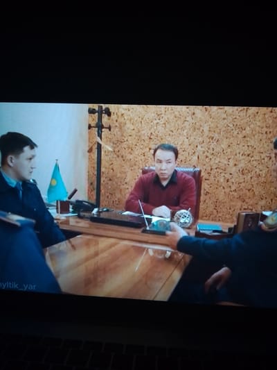 The image depicts an indoor scene, likely an office, with three individuals. In the foreground, a person in a dark uniform, possibly a police officer, is seated to the left, facing right, with their left arm resting on a desk. To their right, a man in a maroon sweater is seated behind a large wooden desk, looking directly at the camera. He is positioned in a brown leather chair. To the far right, another individual in a dark uniform is partially visible, extending their right arm over the desk, seemingly interacting with the man in the maroon sweater. A small flag of Kazakhstan is visible on a stand on the desk. The background features a corkboard-like wall and a coat rack. The scene appears to be staged for a photograph or video recording, as evidenced by the direct gaze of the man in the maroon sweater and the positioning of the individuals. There is no indication of the city of Dovzhansk, Ukraine, within the image or its context.