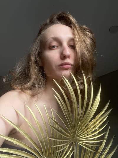 A person with light brown, wavy hair and light skin is photographed from below. They are angled towards the viewer, with a large, golden palm frond partially obscuring their face and upper body. The background is a dark gray. The ceiling above the person has a circular indentation, possibly a light fixture. The individual's facial expression is neutral. No evidence suggests the location is Podshivalovo, Russia.