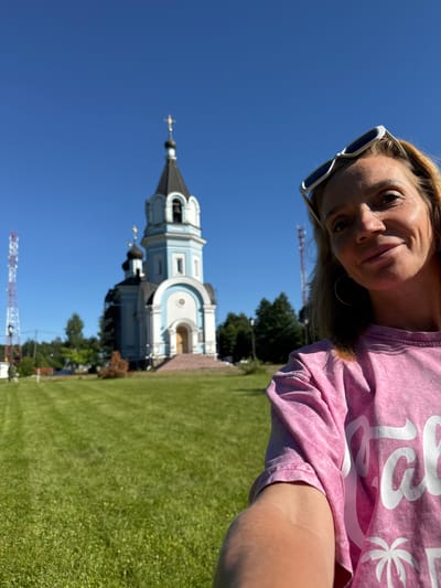 A selfie is taken by a blonde woman with sunglasses perched on her head, smiling at the camera. She wears a pink t-shirt. In the background, a light blue and white orthodox church with a tall steeple stands under a clear blue sky. The church has a black dome with a golden cross on top. In front of the church, a manicured green lawn stretches out. To the left of the church, a tall cell tower with red and white accents is visible. Trees are scattered throughout the scene. The woman appears to be in St. Ozerki, Russia.