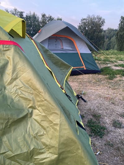 The image shows two tents set up outdoors, likely in a campsite or natural area. The foreground is dominated by a green tent, its fabric wrinkled and folded, with yellow and red trim visible. The tent is positioned in a way that its interior is not visible. In the background, a second tent, gray and green with orange accents, is set up on dry, matted grass. This tent is partially open, revealing a mesh window. The environment appears to be a field with scattered dry vegetation and some patches of green grass. Behind the tents, a line of trees is visible against a pale, overcast sky. The scene suggests an outdoor camping setting, possibly in the vicinity of Peshcherka, Russia, given the context provided, though no explicit city landmarks are visible.