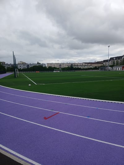 This is a medium-angle, eye-level shot of a purple running track bordering a green soccer field in Charleville-Mézières, France. The perspective is from the side, looking across the field. The sky is overcast with gray clouds, suggesting a daytime scene with diffused light.
On the left side of the image, the green soccer field is visible with white lines marking its boundaries and center circle. A soccer ball rests near the center of the field. In the background, there are rows of multistory buildings with architectural styles indicative of European urban environments. Trees are interspersed between the buildings and along the field's edge. A tall light pole stands prominently in the distance.
The foreground is dominated by the vibrant purple running track, divided by white lane lines. Small blue markers are visible on some of the lanes, and a red marker is also present on one of the lanes. The texture of the track surface is apparent. The edge of the track, where it meets a darker surface, is also visible on the right side of the frame.
There is no visible text in the image. The scene depicts an athletic facility, likely a stadium or sports complex, set within a town or city. The overall mood is calm, with no people or specific events captured, suggesting a moment between activities or before a game.