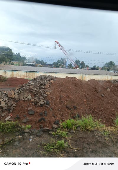 The image depicts an industrial or construction site within Sigona ward, Kenya, under a uniformly overcast sky. In the foreground, large, irregular mounds of reddish-brown soil are interspersed with dark grey rock fragments. Sparse green grass grows from muddy ground at the bottom edge, and a white plastic bottle lies horizontally on the ground near the soil pile. The midground features a long, low-rise building with a light-colored lower wall, displaying dark markings, and a dark, corrugated roof, secured by razor wire along its top. Behind this building, an industrial complex is visible, containing yellow heavy machinery and other structures. A prominent red lattice-boom crane with a dual-hook hoist system is positioned centrally in the midground, its boom extending upwards and to the right. Multiple parallel utility lines traverse horizontally across the mid-to-upper frame. A dense line of green trees flanks the left side and extends into the background, where additional structures, including a water tower, are partially obscured by atmospheric conditions. No individuals are visible, and no active human interactions are depicted.