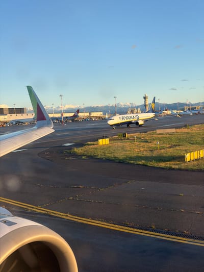 The image captures an airport scene from the perspective of an aircraft window, looking down onto the tarmac. The perspective suggests the viewer is aboard a plane, as a portion of its wing, featuring a green and red wingtip, and the engine nacelle are visible in the right foreground.

In the midground, a white Ryanair Boeing 737 aircraft is prominent, either taxiing or parked on the asphalt tarmac. The word "RYANAIR" is clearly visible in blue lettering on its fuselage. The tarmac features yellow painted lines and markings, as well as drainage grates. Adjacent to the tarmac are green grass verges and yellow informational signs, one of which shows the letter "W". Other aircraft are visible further in the distance, including one partially showing "cargo" on its livery.

The background reveals a clear blue sky with a few faint clouds. In the far distance, a range of snow-capped mountains stretches across the horizon, indicating a mountainous geographical setting. The lighting, characterized by warm tones and long shadows, suggests it is either early morning or late afternoon (golden hour).

The overall scene depicts typical airport activity on a clear day. Given the location context of Lonate Pozzolo, Italy, and the visible snow-capped mountains, this airport is likely Milan Malpensa Airport (MXP), which is situated near the Alps.