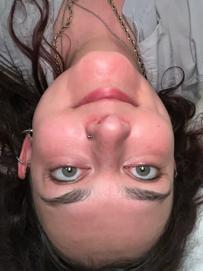 A close-up, angled shot of a person's face, featuring their green eyes and a nose piercing. The person has dark hair. Gold chains are visible around their neck and the person is wearing a light gray shirt. The location in the city of Puçol, Spain is not discernible from the visual information provided.