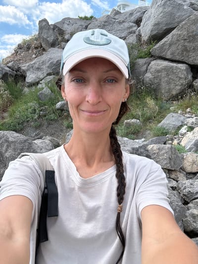 A woman with blue eyes and a light blue cap with a smiling face logo is looking at the camera. She has a braid of dark brown hair that reaches her shoulder. She is wearing a light-colored t-shirt and a grey backpack. The background consists of large grey rocks and some green vegetation, with a bright blue sky and white clouds overhead. A white building is visible on a rocky hilltop in the distance. The woman appears to be outdoors in a rocky, possibly mountainous area, near a coastal town, or a rocky coastline. The location is not Bondarenkovo, Ukraine.