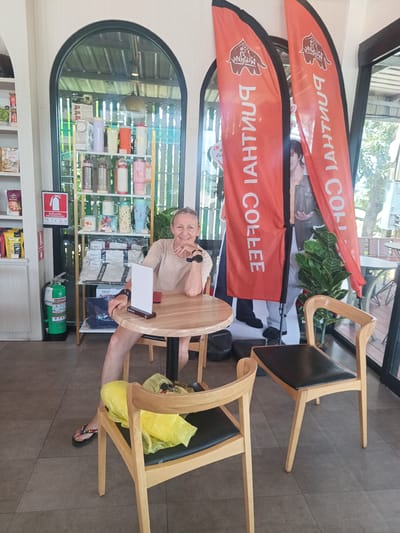The image depicts a woman seated at a small, round wooden table in a coffee shop in Plai Phraya, Thailand. The coffee shop appears to be an outlet of "Puritai Coffee," as indicated by the orange banners displaying the company's logo and name, which are positioned near the window to the right. The woman is fair-skinned with short blonde hair and is wearing a tan-colored shirt. She rests her right elbow on the table and is smiling. A small, white menu stand is placed on the table.

The foreground contains two wooden chairs with black seats. The chair on the left holds a yellow plastic bag containing several items. Behind the woman is a shelving unit stocked with various merchandise, including thermoses, mugs, and packaged goods. A red fire extinguisher is visible near the shelving unit. Behind the banners is a life-size standee of a person, obscured in part by foliage. Outside the coffee shop, greenery and a glimpse of the surroundings can be seen. The floor is tiled in a dark gray color.