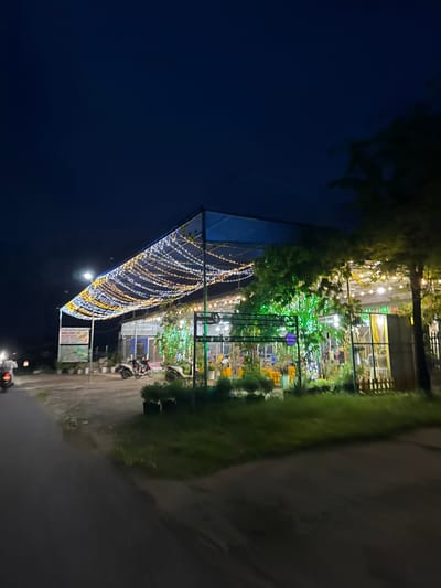 In the city of Hàm Thuận Bắc, Vietnam, at nighttime, a building with a blue tarp roof is illuminated by a festive string of lights. The lights are arranged in a dense, undulating pattern, creating a canopy effect over an outdoor seating area. The building appears to be a restaurant or cafe, with tables and chairs visible under the lights. Green lights adorn a nearby tree, adding to the vibrant atmosphere. Two motorcycles are parked outside the building. The scene is bordered by a dark road in the foreground and the dark night sky above.