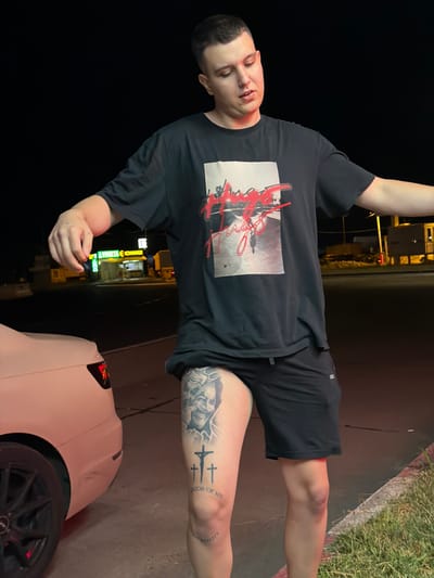 A young man stands in front of a light-colored sedan at night. He is wearing a black t-shirt with a graphic print and black shorts. He has a tattoo on his right thigh of a joker's face and three crosses with the words "ATOR OF MY REALITY" below them. The man is looking down and to his right, with his arms slightly extended outwards. In the background, there is a brightly lit store with a sign that reads "VINETTA EXCHANGE". The scene appears to be a street in the city of Giurgiu, Romania.