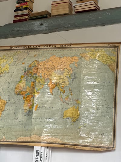 The image shows a wall in what appears to be a classroom or library. A large, old, pull-down world map, labeled "ПОЛИТИЧЕСКАЯ КАРТА МИРА" (Political Map of the World), is prominently displayed. The map is encased in clear plastic, which has some wrinkles and distortions. Above the map, a rough, light blue wooden beam runs horizontally across the image. Stacked on this beam are several books, mostly with red, tan, and dark covers. Some of the books have Cyrillic lettering visible on their spines. Below the map, a portion of a roll of paper or similar material is visible, with the word "APER" printed on it multiple times. The lighting is somewhat dim, suggesting an indoor setting. The context provided suggests this location is in Kuybyshev, Russia, though this cannot be visually confirmed from the image alone. There are no people visible in the image.