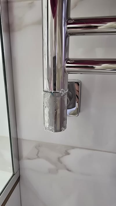 The image shows a close-up of a chrome heated towel rail mounted on a white marble-patterned tiled wall in a bathroom. The lower portion of the vertical bar of the towel rail is covered in a thick, white, crystalline buildup, possibly limescale or mineral deposits. The buildup is uneven and appears to be dripping down the bar. A square chrome mounting bracket is visible behind the lower part of the towel rail. To the left, a sliver of a mirrored cabinet or shower enclosure can be seen. The overall impression is one of a bathroom fixture in need of cleaning. There are no people or animals present. The location is not specified in the image itself, and the request to include the city of Kamenka, Russia, is extraneous to the visual information provided.