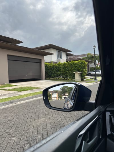 The image shows the side view mirror of a car parked in a residential neighborhood in Río Oro, Costa Rica. The mirror is reflecting a street with several houses and trees. The driver's side window is visible. The car is parked on a cobblestone driveway in front of a two-story house with a closed garage door. There are several green bushes in front of the house. The street is empty. The sky is cloudy. The coordinates, latitude: 9.945, longitude: -84.201, place the location in a suburb of San José, Costa Rica.