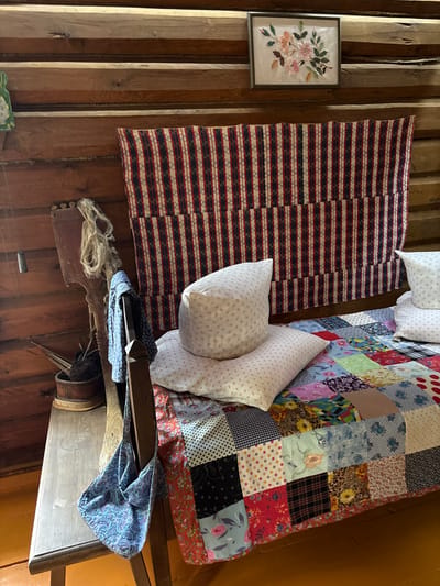 The image depicts a rustic bedroom, likely in a rural setting or a historic home. The walls are made of dark, horizontally aligned logs. A large, colorful patchwork quilt covers a bed, with several pillows arranged on top. Above the bed, a striped textile hangs vertically. To the left of the bed, there is a wooden bench with a patterned fabric bag draped over its edge. Various rustic tools and materials, such as a wooden paddle and a ball of yarn, are also visible on the bench. A framed embroidery of flowers hangs on the wall above the striped textile. The overall impression is one of a traditional, cozy interior, possibly in the village of Ovsyanka, Russia, known for its association with writer Viktor Astafyev and its preserved rural character.